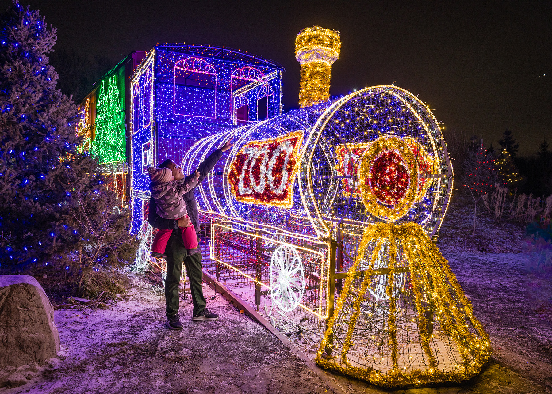 A dad holds his child up to look at a life light train made of lights